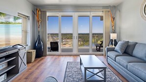 A spacious living room with a large sliding glass door leading to a balcony, a flat-screen TV, a grey sofa, and a coffee table. - Luxury and Spacious Condo With Spectacular View (Panama City Beach)