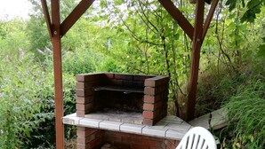 An outdoor brick grill under a wooden pergola, surrounded by greenery.