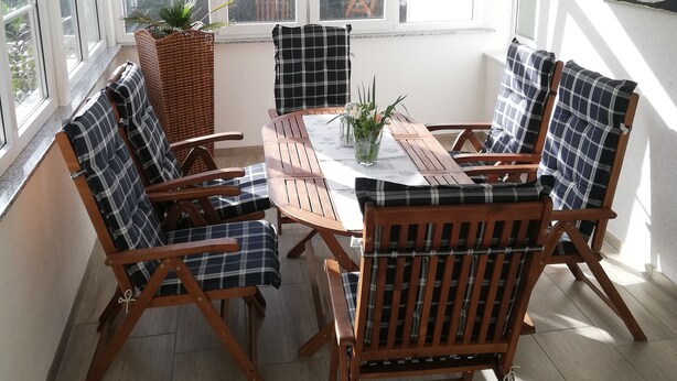 A sunlit room with a round wooden table, six plaid chairs, and a large window with a view of trees.