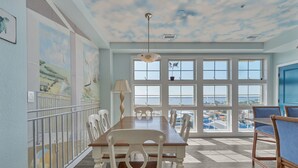 A dining area with a long table, white chairs, and a view through large windows. - Book Sandtastic Penthouse 421A | Sandbridge Vacation Rental (Virginia Beach)