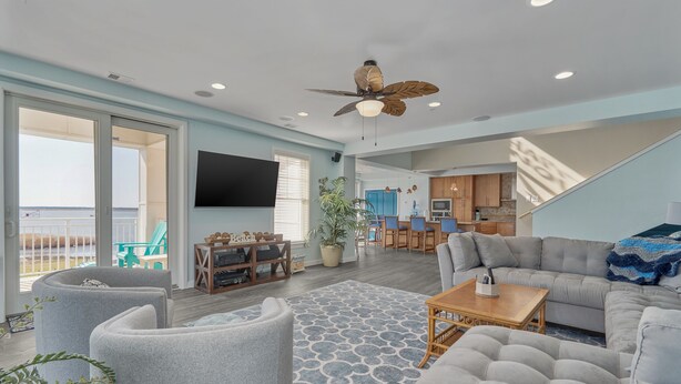A spacious living room with a large grey sectional sofa, a grey armchair, and a wooden coffee table. There is a ceiling fan, a flat-screen TV mounted on the wall, and a potted plant. - Book Sandtastic Penthouse 421A | Sandbridge Vacation Rental (Virginia Beach)