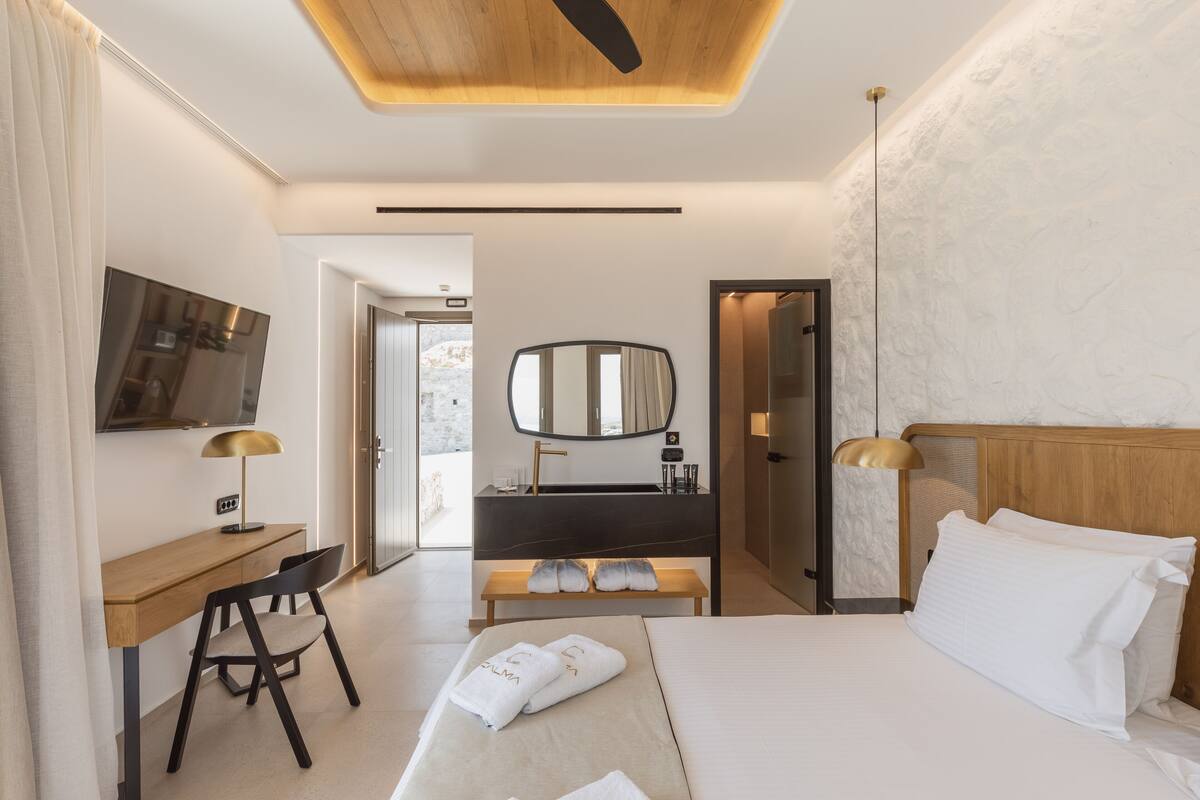 A modern hotel room with a wooden ceiling, a large bed, a desk with a chair, a vanity with a mirror, and a television on the wall.