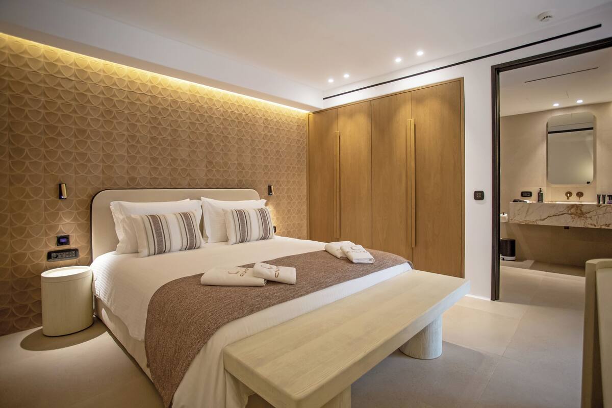 A modern hotel room with a large bed, a bedside table, a bench, a nightstand, and a bathroom visible through an open door.