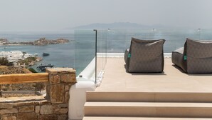 A terrace with two cushioned chairs and a glass balustrade overlooking a coastal area with boats and buildings. - Calma Suites Mykonos (Mykonos)