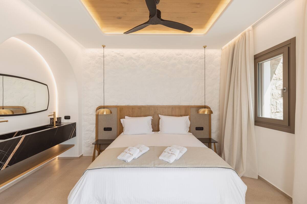 A modern hotel room with a large bed, a wooden ceiling fan, a built-in TV, and a large window with curtains.