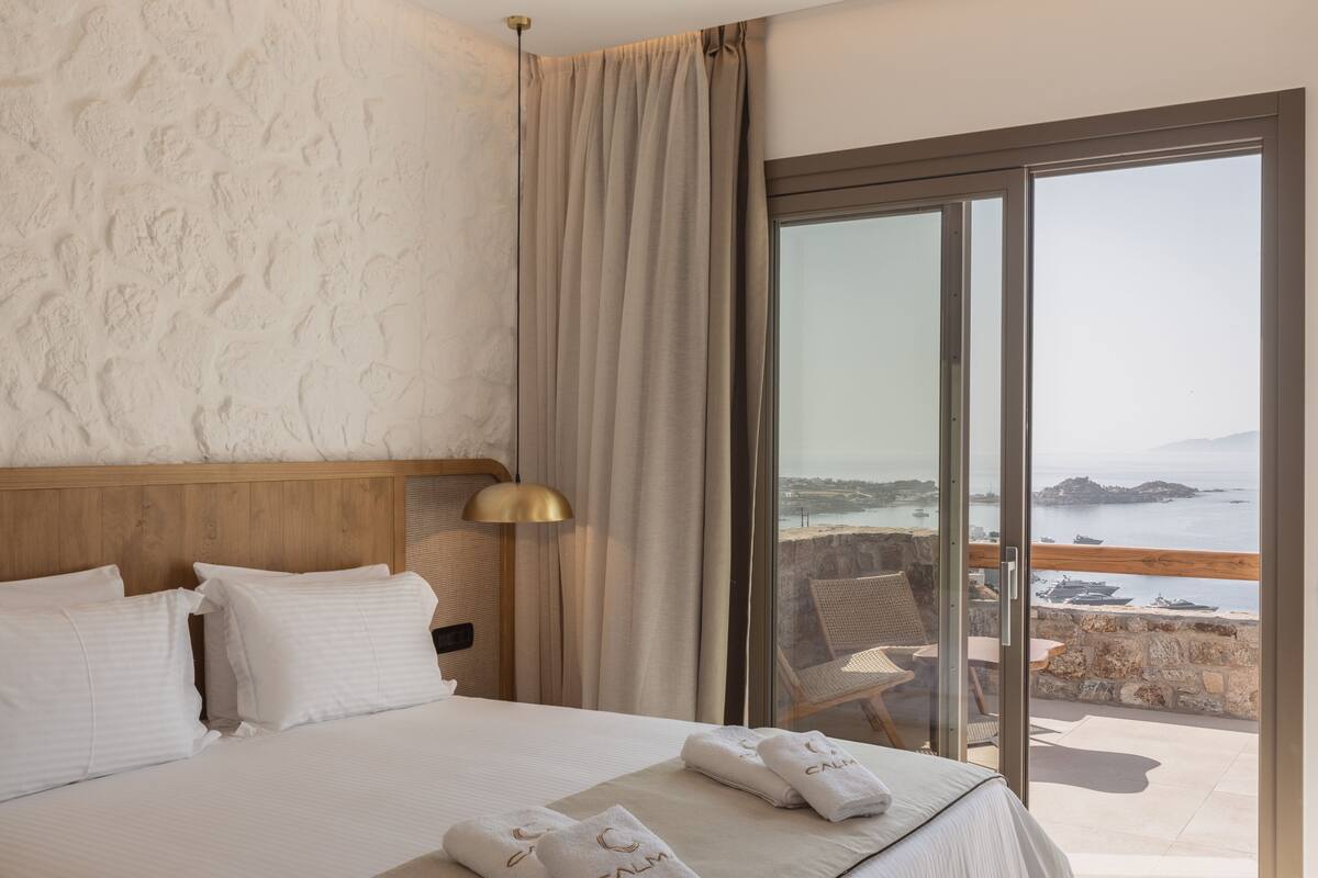 A hotel room with a bed, a large window with a view of the sea, a wooden headboard, and a nightstand with a lamp.