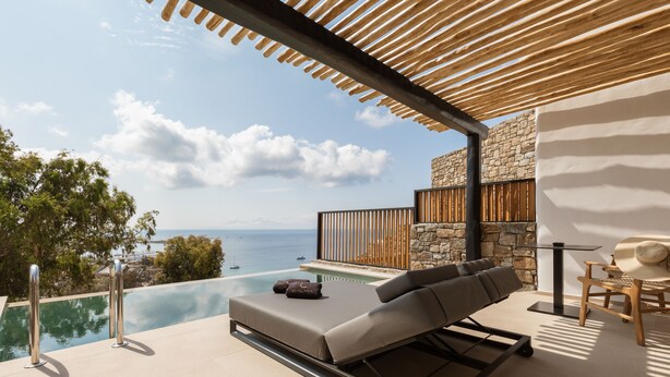 A modern outdoor lounge area with a pool, a stone wall, and a view of the sea. - Calma Suites Mykonos (Mykonos)