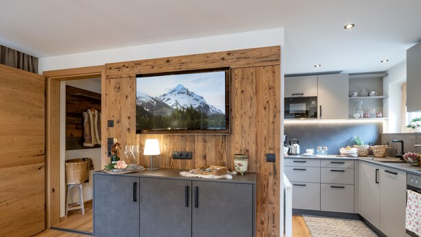 A modern kitchen with wooden cabinetry, a large flat-screen TV mounted above the counter, and a wooden-framed wall. - Landhaus Dengg in Gerlos: Rosengarten Apartment for 2 to 4 people (Gerlos)