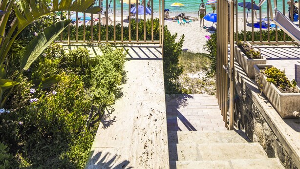 Eine Steintreppe am Strand, die zu einem sandigen Bereich mit Sonnenschirmen und Personen führt.