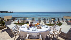 Ein Esstisch mit Frühstücksaufmachung, der Blick auf das Meer bietet, umgeben von Stühlen und einem Geländer mit Ausblick auf den Strand.