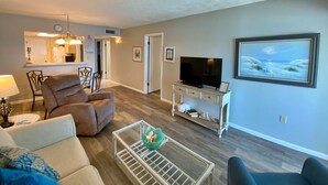 A living room with a sofa, a coffee table, a TV on a stand, and a painting on the wall. - Beachfront, Pool & Sunroom (Gulf Shores)