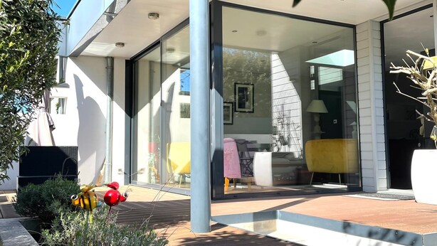 A modern house with a glass door entrance, a patio with outdoor furniture, and a garden with decorative plants.