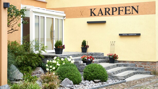 A hotel entrance with a yellow facade, steps leading up to the door, and potted plants. - Hotel-Restaurant Zum Karpfen (Obernburg)