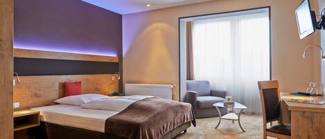 A hotel room with a large bed, a desk, a chair, a small table, and a TV mounted on the wall.