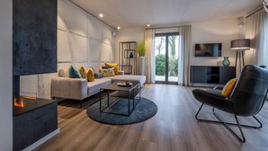 A modern living room with a sofa, coffee table, and a black leather chair. There is a fireplace, a TV, and a large window with curtains. - Büchelbergerei Chaletdorf (Gunzenhausen)