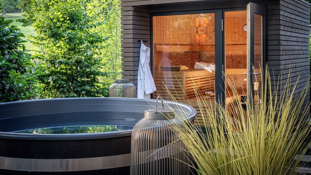 A modern outdoor sauna with a wooden exterior and glass walls, surrounded by lush greenery. - Büchelbergerei Chaletdorf (Gunzenhausen)