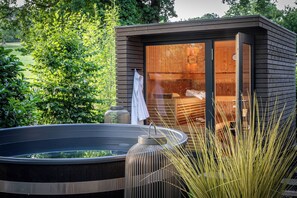 A modern outdoor sauna with a wooden exterior and glass walls, surrounded by lush greenery. - Büchelbergerei Chaletdorf (Gunzenhausen)