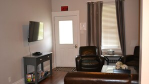 A living room with a flat-screen TV, a leather armchair, a sofa, a small table with decorative items, and a bookshelf. - 5 bed 2 Bath in Downtown Hermann, MO (Hermann)