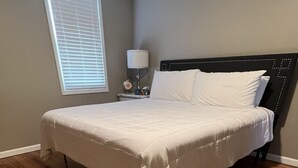 A bedroom with a large bed, wooden flooring, a nightstand with a lamp, and a window with blinds. - 5 bed 2 Bath in Downtown Hermann, MO (Hermann)