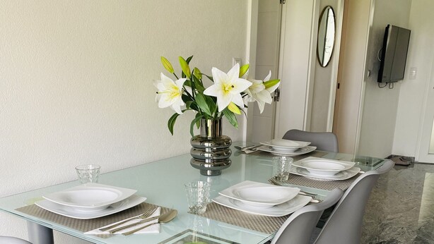 Un comedor con una mesa de vidrio, sillas grises y un florero con flores.