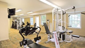 A gym with various exercise machines, a television mounted on the wall, and large windows providing natural light.