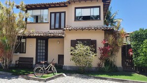 Uma casa de dois andares com telhado de cerâmica, uma bicicleta estacionada em frente e um jardim bem cuidado com diversas plantas.