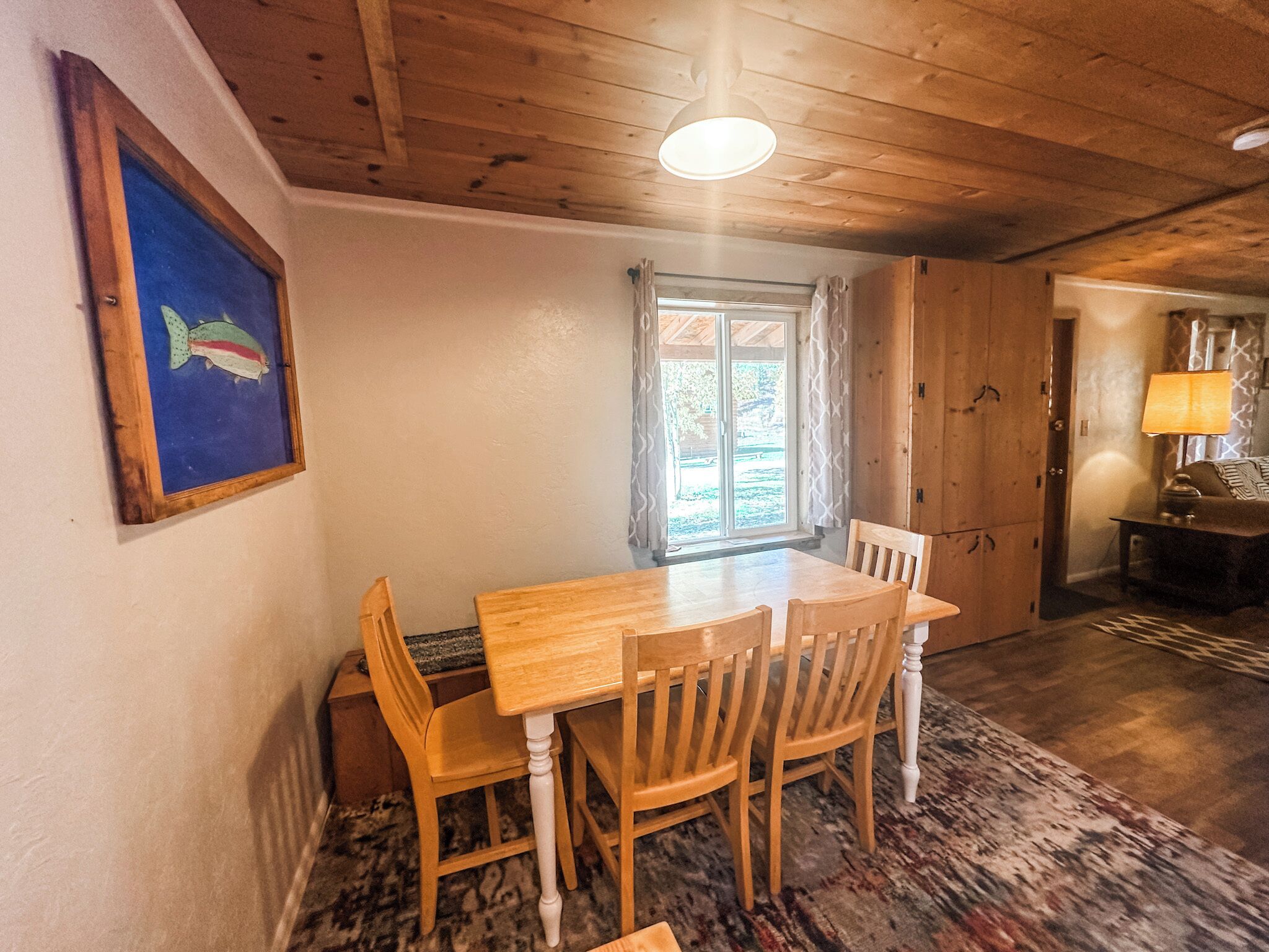 A wooden dining table with chairs, a framed fish painting, a window with curtains, and a lamp on a side table.