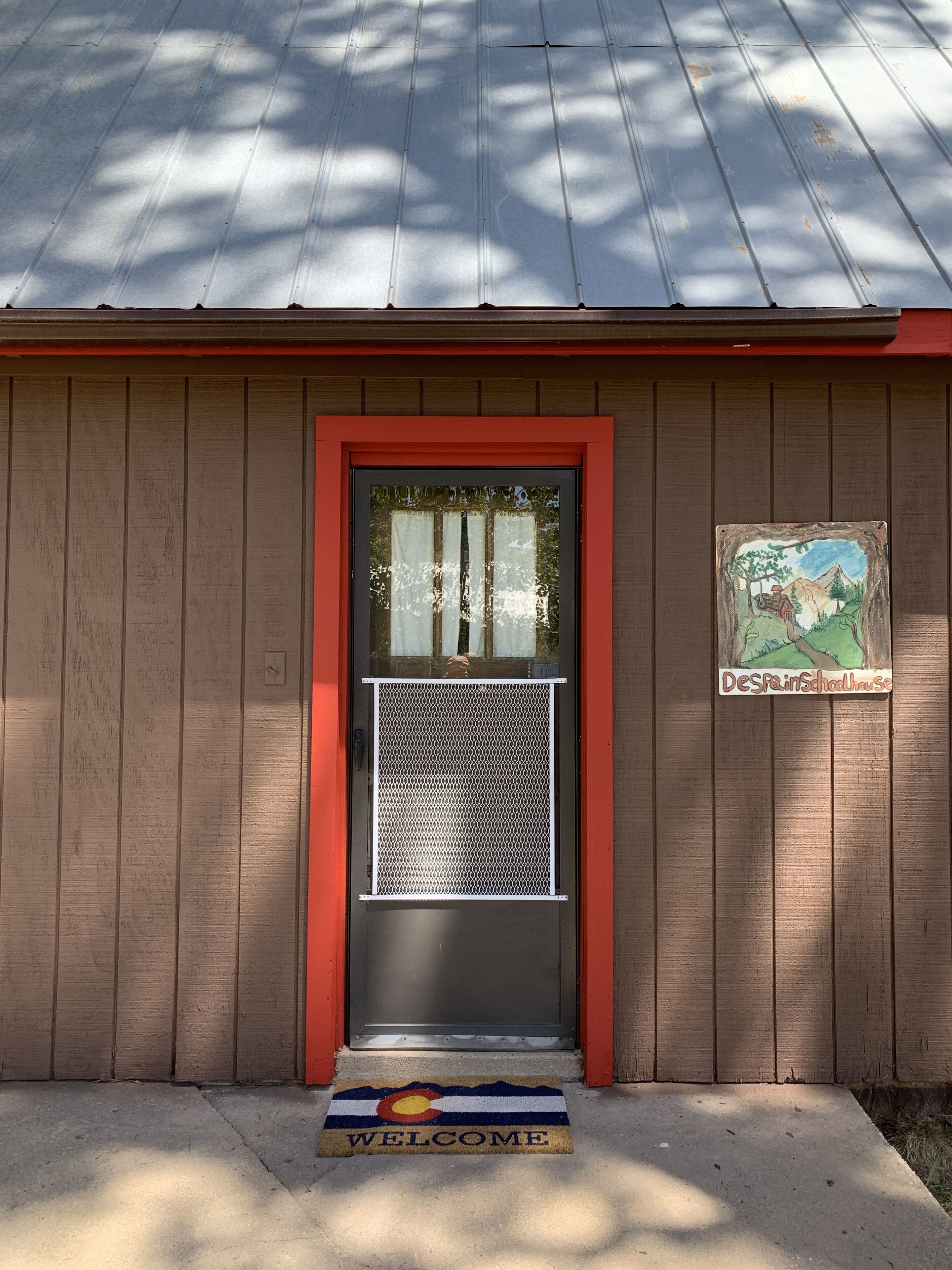A wooden building with a red-framed door, a mesh screen, and a welcome mat with a state flag.