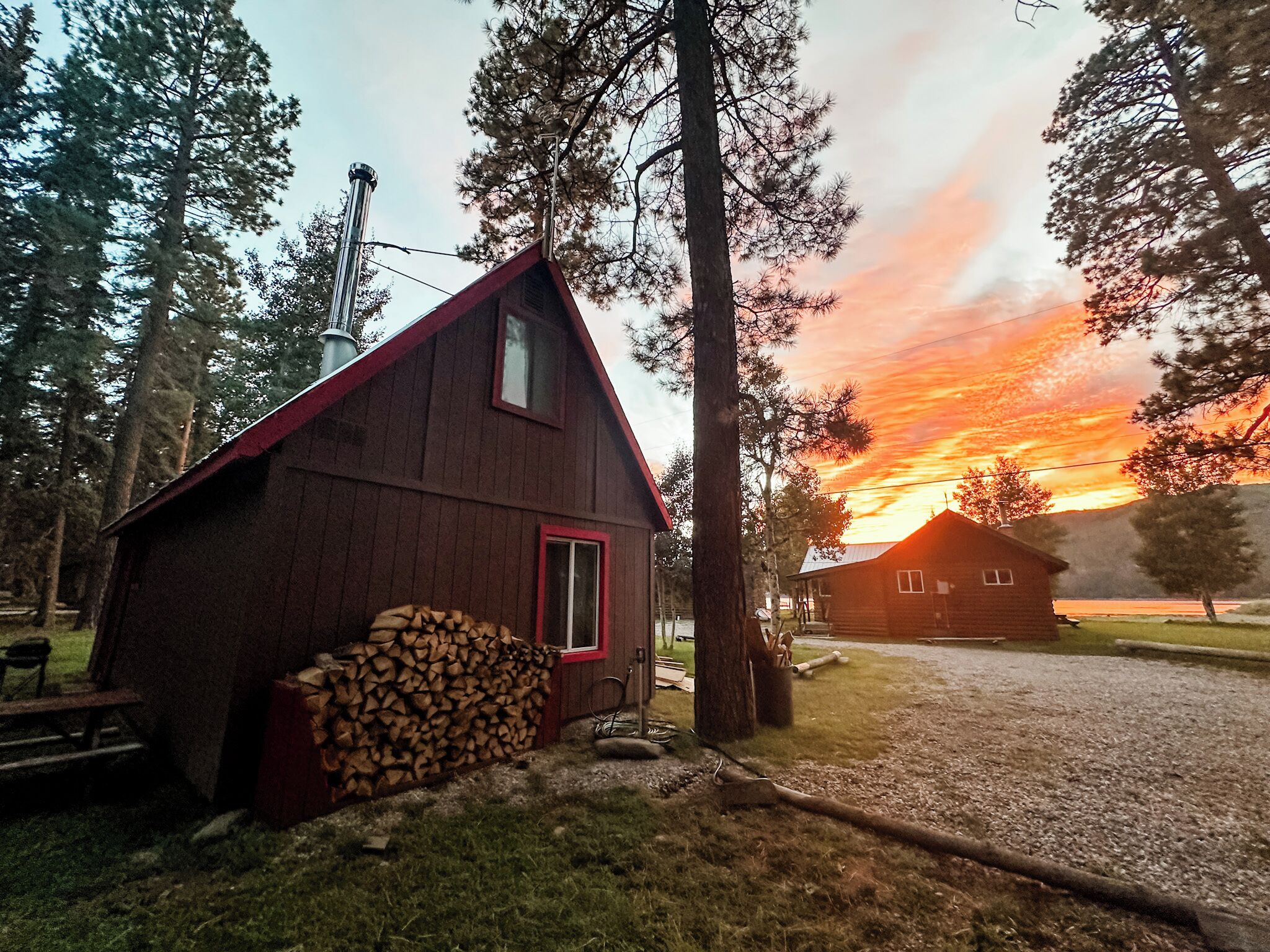Un chalet rustique avec un toit rouge et une cheminée, entouré d’arbres et bordé d’un chemin de gravier.