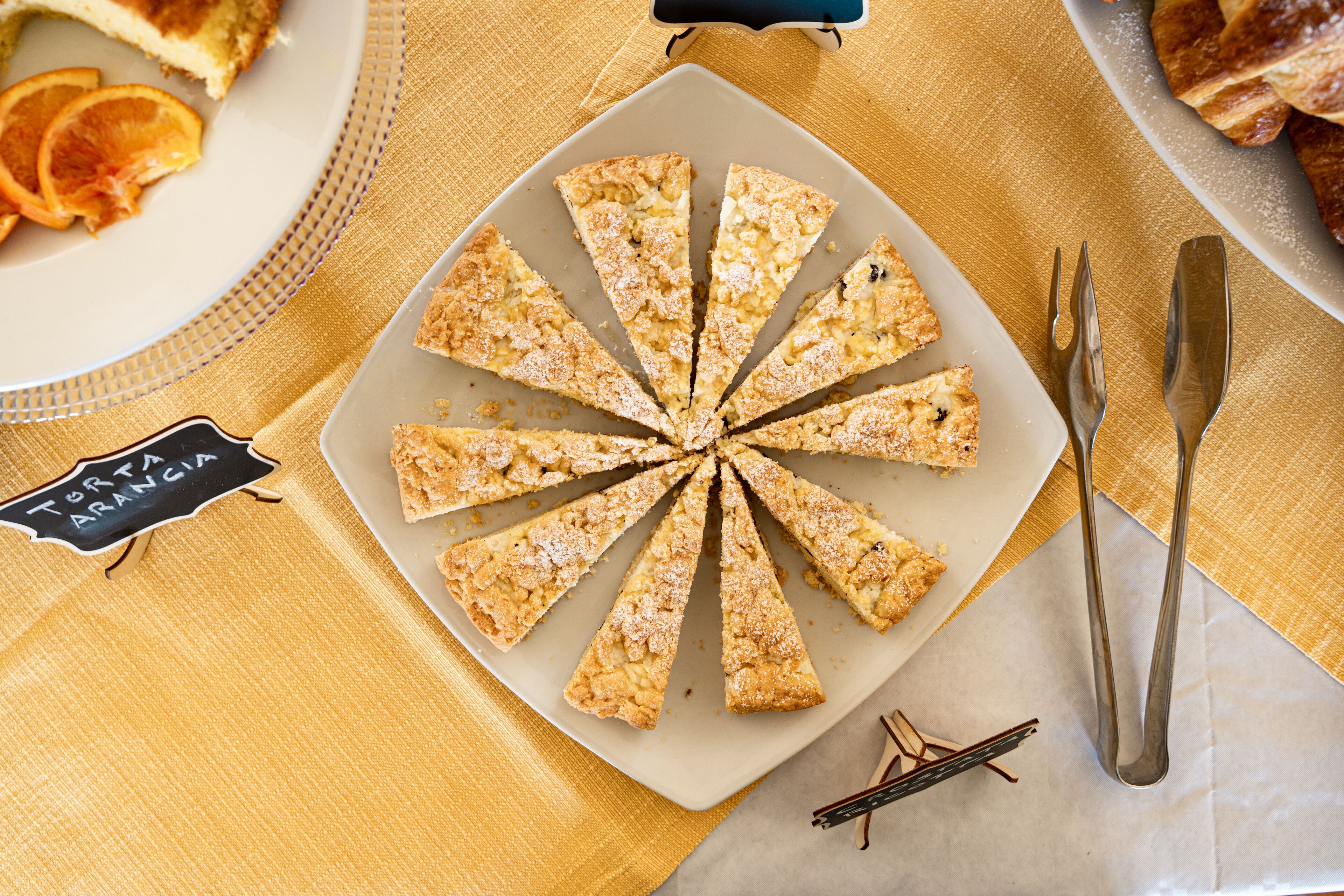 A plate of triangular pastries with a sign reading "Torta Arancia" on a table with a yellow tablecloth.