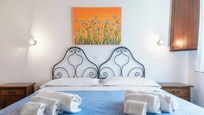 Una cama cuidadosamente hecha con una colcha azul estampada y almohadas blancas, dos toallas blancas enrolladas sobre la cama, un cuadro en la pared con flores azules sobre fondo naranja, y mesitas de noche de madera con apliques.