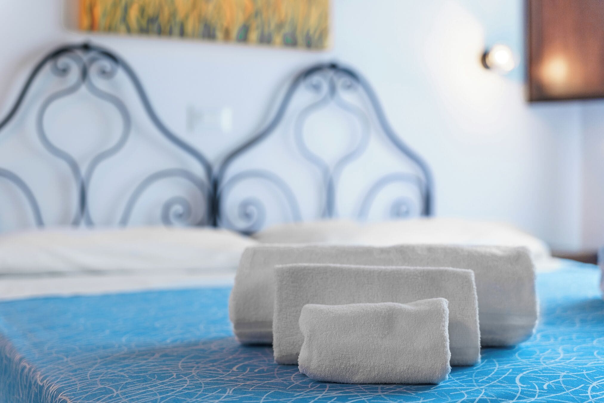 A bed with a blue textured cover, two rolled towels, and a metal headboard.