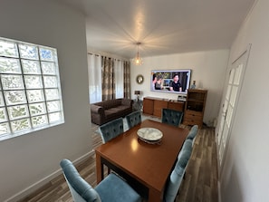 A dining area with a wooden table, blue upholstered chairs, a television mounted on the wall, and a sofa in the background.