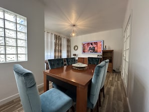 A dining area with a wooden table, blue upholstered chairs, a chandelier, a television, and a window with a view of the outdoors.