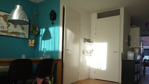 Een kamer met een houten vloer, een bureau met stoelen, een hanglamp, een wereldkaart, een klok en een deur met een raam.