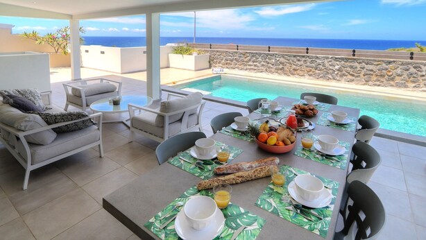 A dining area with a view of the ocean, a pool, and a stone wall.