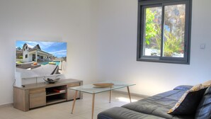 A modern living room with a glass-top coffee table, a leather sofa, a flat-screen TV, and a window with a view of a house and greenery.