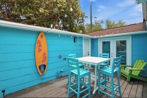 Une maison bleue avec une planche de surf accrochée au mur, une table avec des chaises, et une chaise verte à l’extérieur.