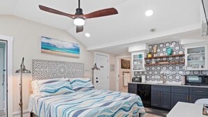 A bedroom with a bed, a ceiling fan, a nightstand, a desk, and a kitchen area with cabinets and a countertop.