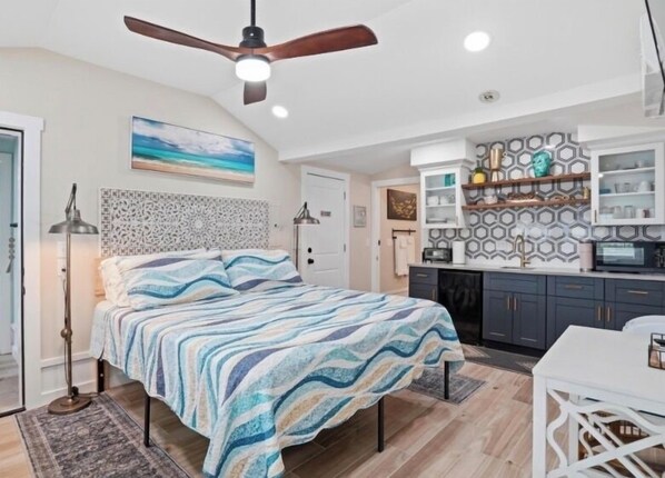 Une chambre comprenant un lit, un ventilateur de plafond, une table de chevet, un bureau et un coin cuisine avec des placards et un plan de travail.