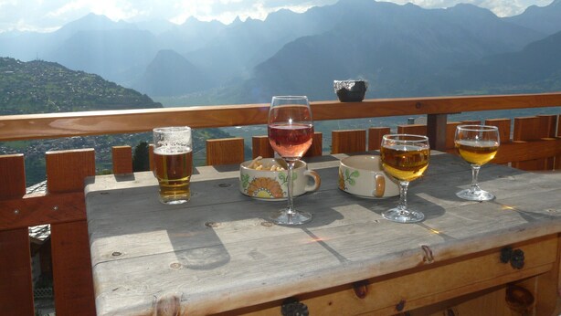 Una mesa de madera con cuatro copas de vino y un bol de frutos secos, con vistas a un paisaje montañoso.
