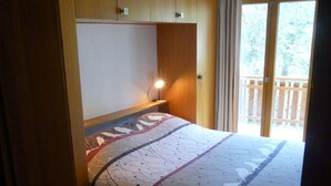Un dormitorio con techo de madera, una cama con colcha estampada, una lámpara de noche y vistas a un balcón a través de una puerta corrediza.