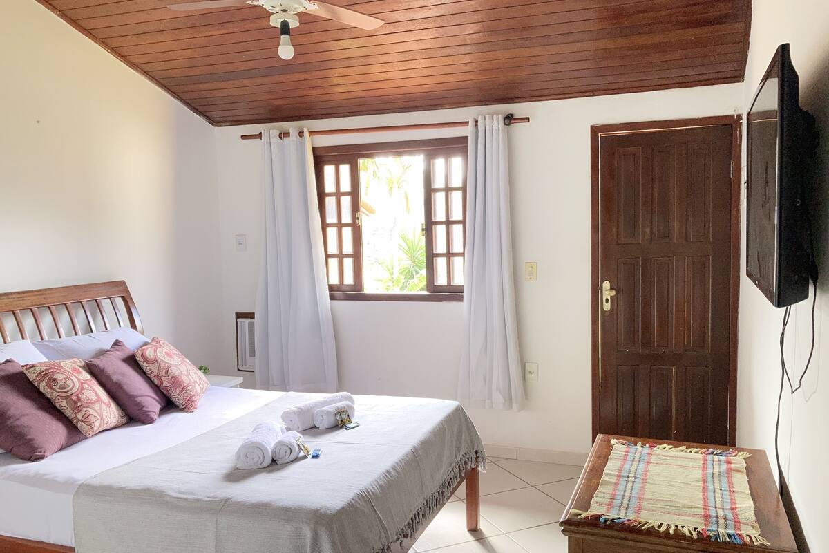Un dormitorio con techo de madera, una cama con cubrecama blanco y toallas enrolladas, una mesita con mantel escocés y una puerta de madera.