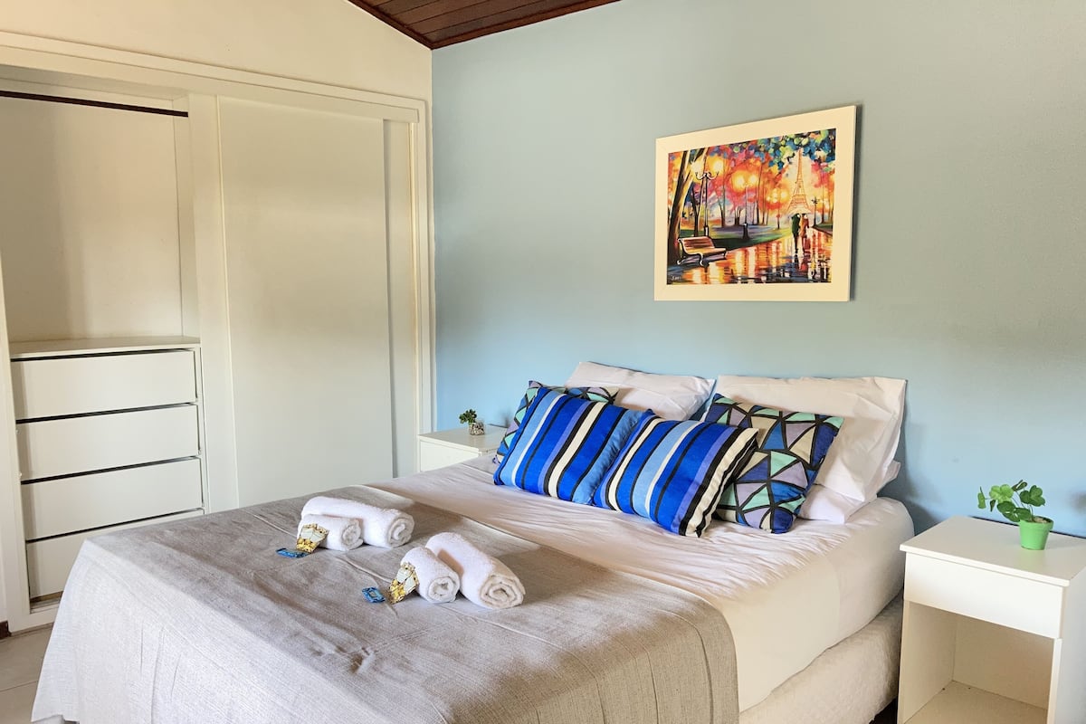 Una cama bien tendida con una manta color beige y almohadas, una mesita de noche blanca con una planta verde, y un cuadro de un paisaje urbano en la pared.