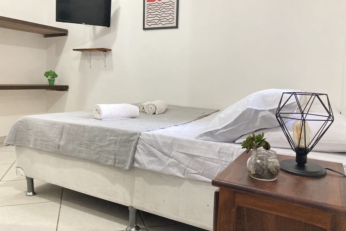 Una cama bien tendida con una manta gris y toallas enrolladas, una pequeña planta en maceta, un frasco de vidrio decorativo sobre una mesita de noche de madera, y una obra de arte enmarcada sobre la cama.