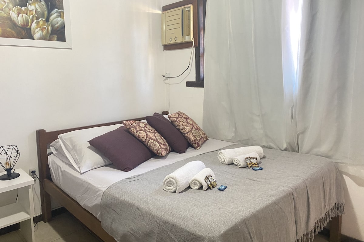 Una cama bien hecha con almohadas blancas y estampadas, una manta gris y toallas enrolladas sobre un cabecero de madera en una habitación con paredes blancas y un cuadro sobre la cama.