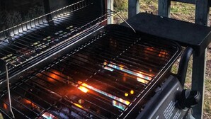 An outdoor grill with a metal grate and a handle, set against a backdrop of trees and a wooden deck. - Trails Head Cabin (Rock Hill)