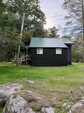 Une petite cabane noire avec un toit en métal, entourée d’une zone herbeuse et d’arbres.