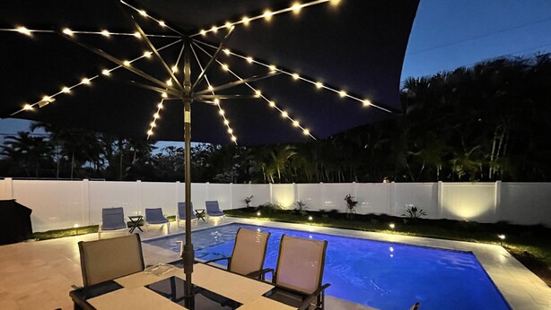 An outdoor pool area with a table and chairs, string lights, and a white fence.