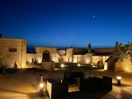 A courtyard with stone buildings, outdoor seating, and a view of the sea at dusk.